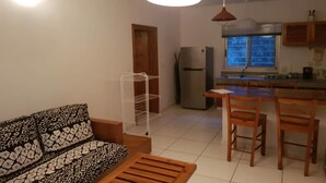 A living room with a patterned sofa, a wooden coffee table, and a white wire shelf unit. There is a kitchen area with wooden chairs and a refrigerator. - Residence Zety (Abidjan)