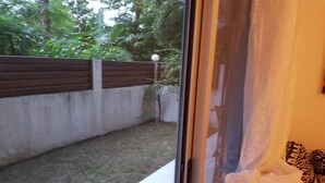 A view through a glass door into a backyard with a wooden fence, a small tree, and a concrete wall. - Residence Zety (Abidjan)