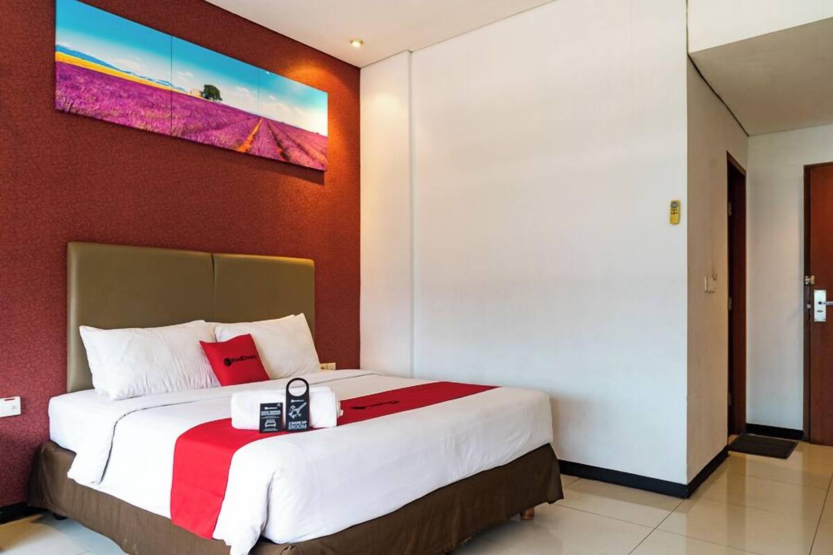 Una habitación de hotel con una cama grande, una colcha roja y blanca, un cabecero y un cuadro de un campo en la pared.