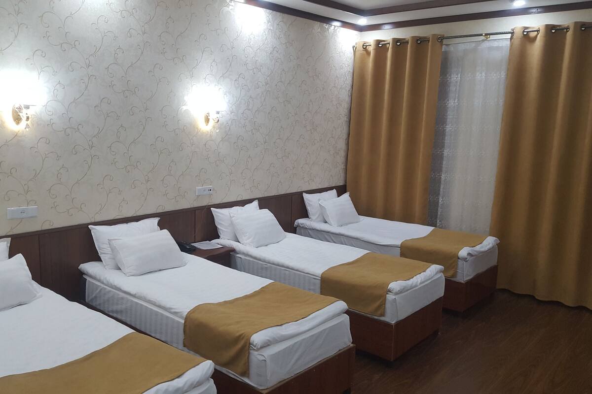 Habitación de hotel con cuatro camas individuales, cada una con ropa de cama blanca y una manta marrón. La habitación tiene papel tapiz estampado y cabecero de madera. Hay dos luces fijadas a la pared y una ventana con cortinas.