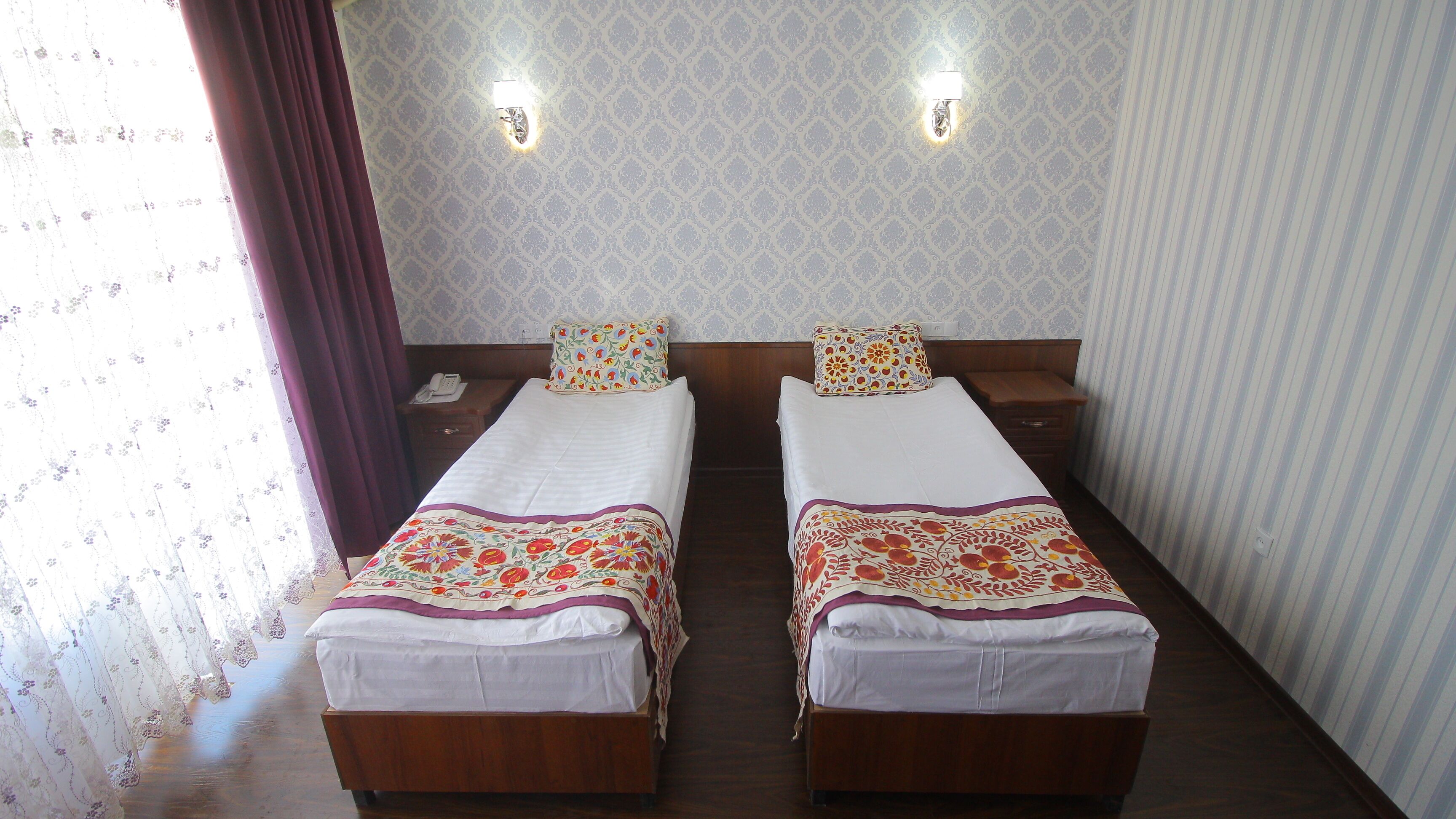 Habitación doble con dos camas, cada una con una colcha y almohadas estampadas con motivos florales. Hay una mesita de noche de madera con un teléfono, y el cuarto tiene papel tapiz decorado y una ventana con cortinas.