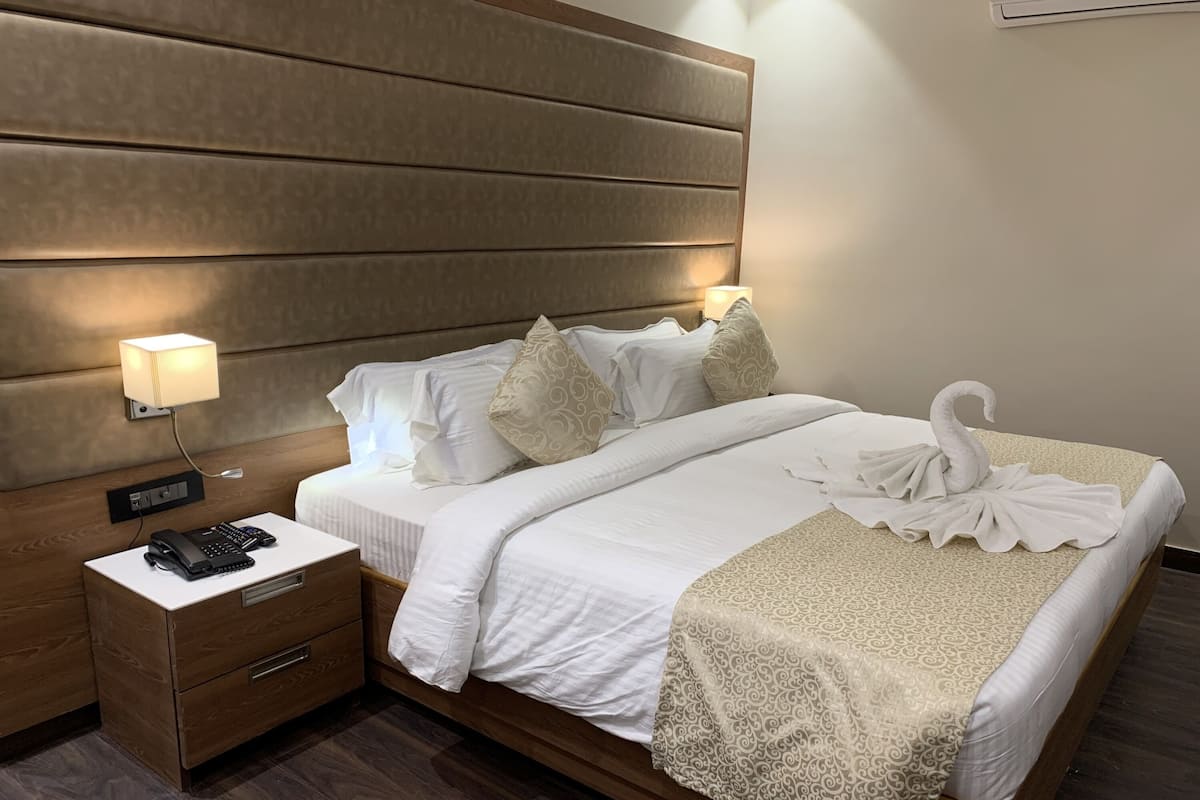 Una cama bien hecha con una toalla con forma de cisne, una lámpara de noche y un teléfono sobre una mesita de madera.
