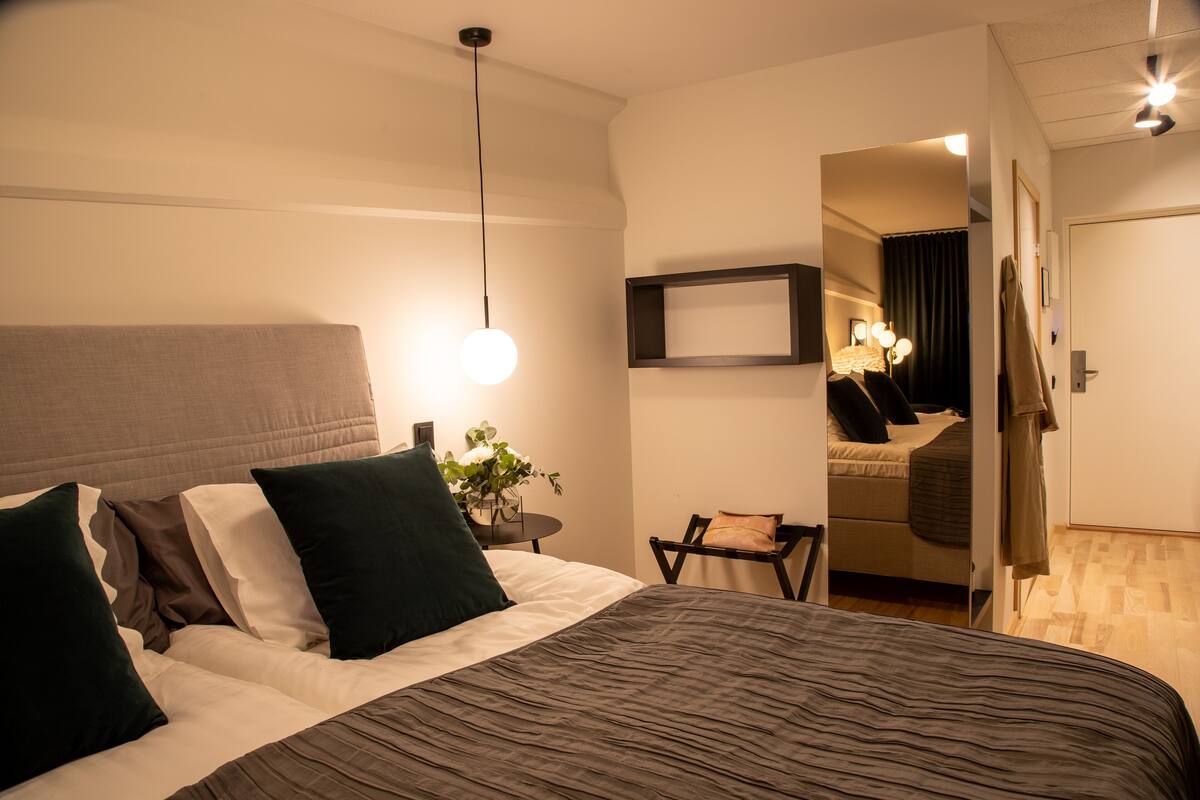 Une chambre d’hôtel moderne avec un grand lit, une table de chevet avec une lampe et un miroir qui reflète la pièce.