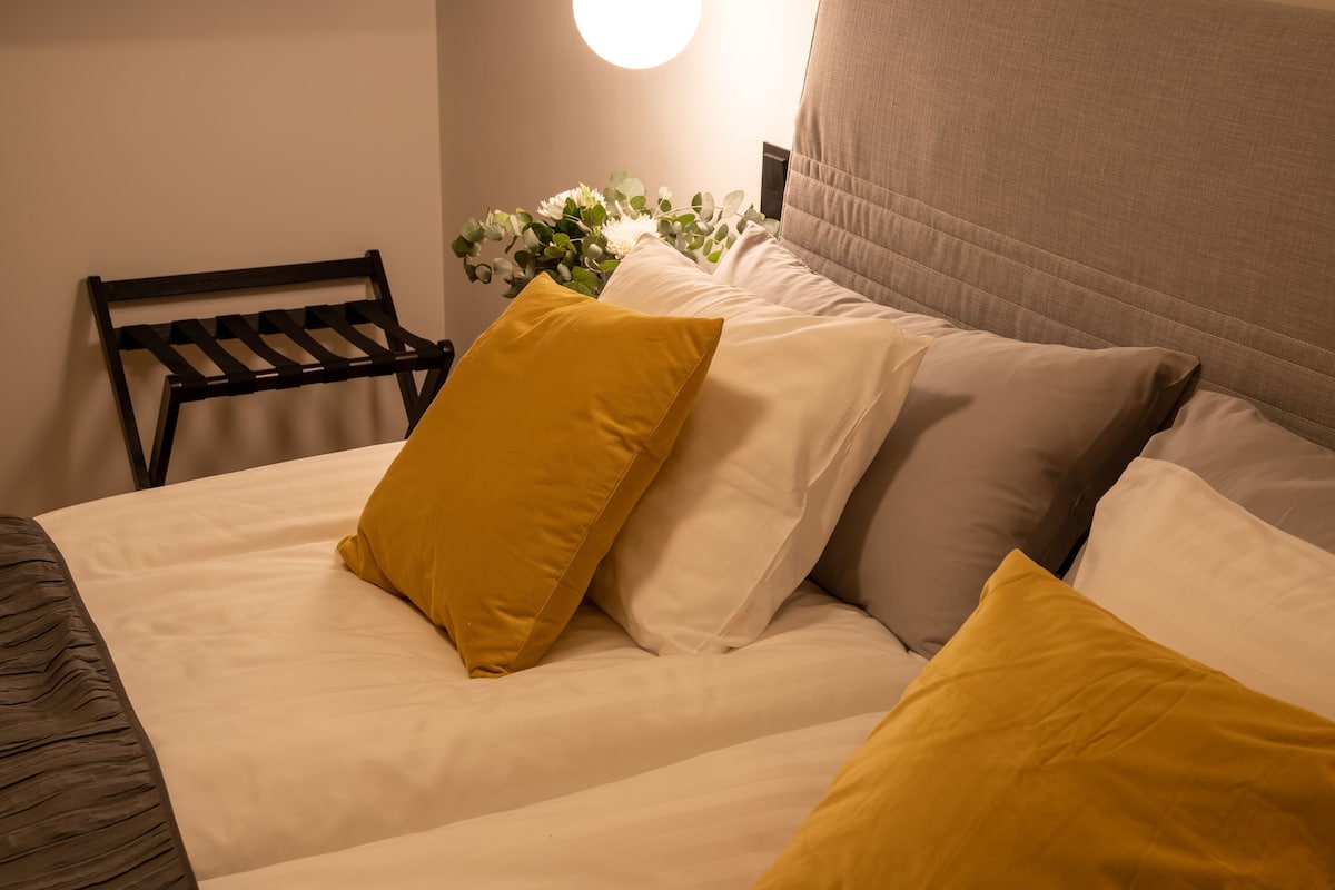 Un lit bien fait, avec des oreillers blancs et jaunes, une tête de lit et un luminaire fixé au mur.