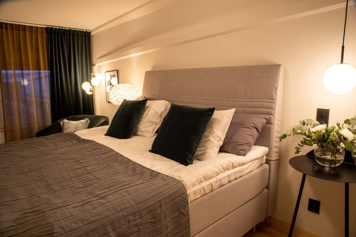 Un lit bien fait, avec une tête de lit, une table de chevet avec une lampe et une fenêtre avec des rideaux.