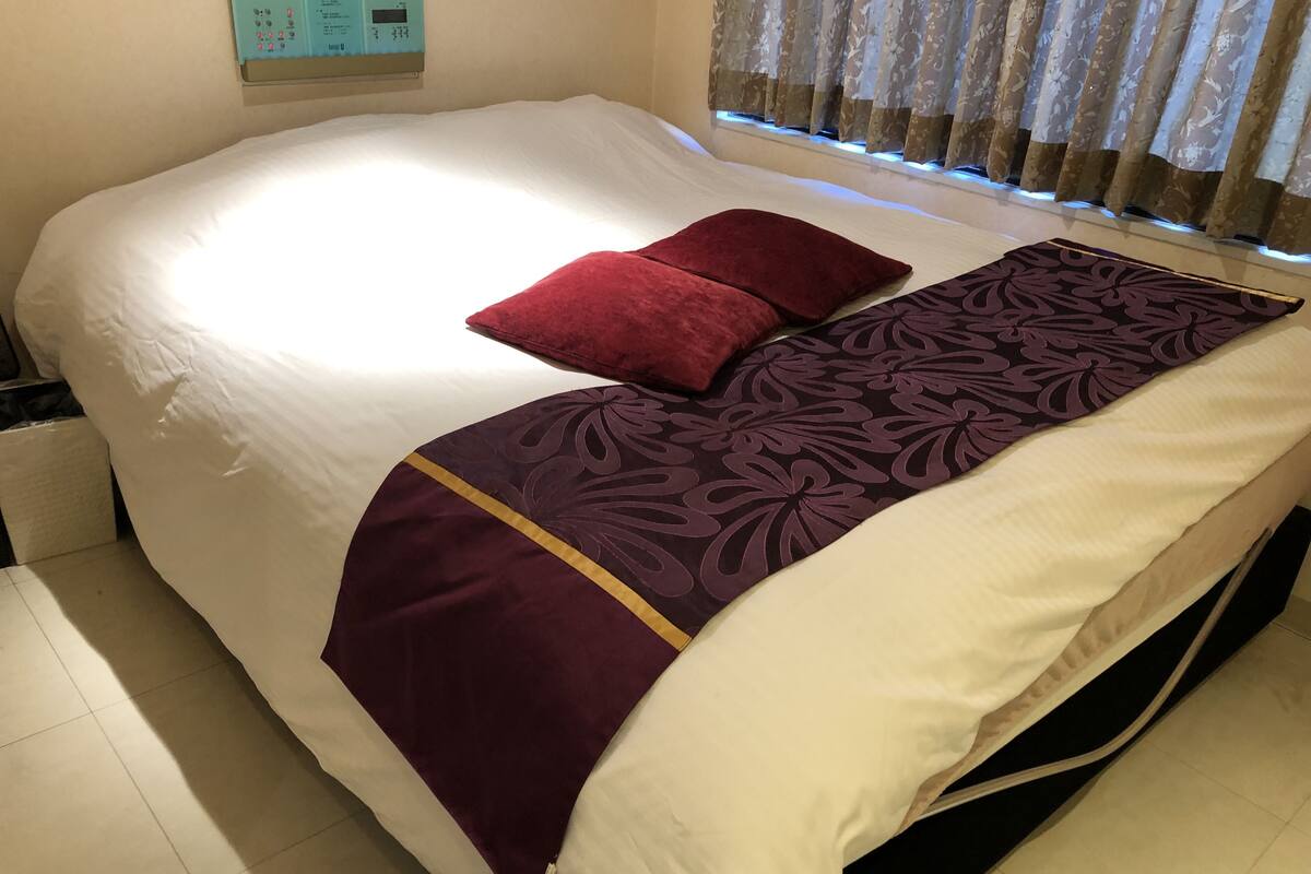 A neatly made bed with white sheets, a purple floral bedspread, and two red pillows.