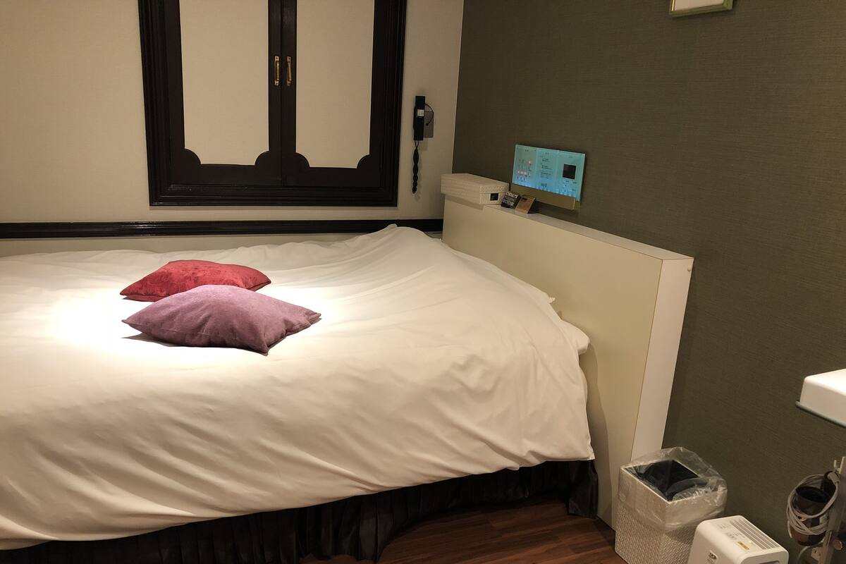 A neatly made bed with white and purple pillows, a wooden floor, a radiator, a trash bin, and a small heater on the floor.