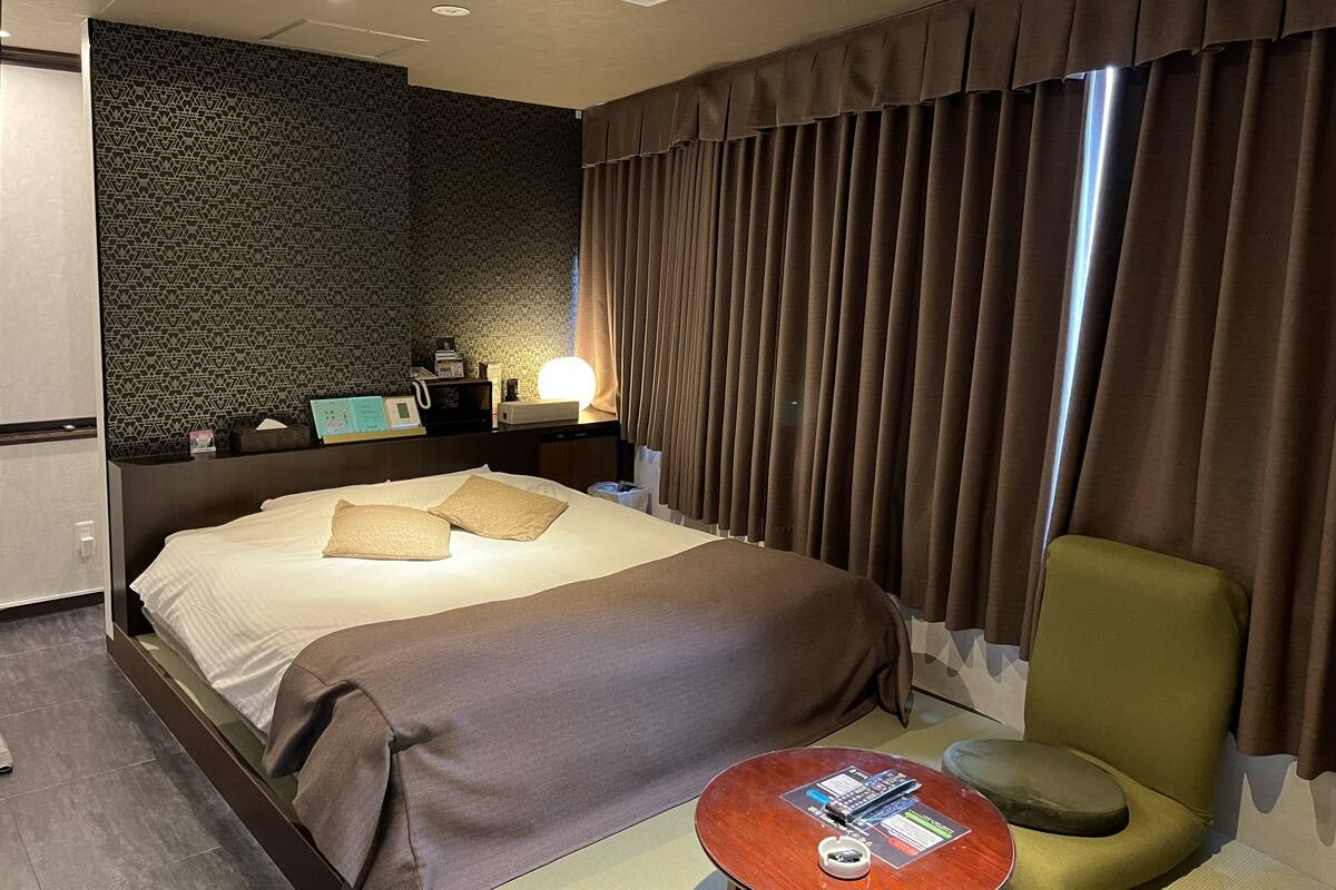 A hotel room with a bed, a bedside table, a lamp, a chair, and a small table with a remote control.