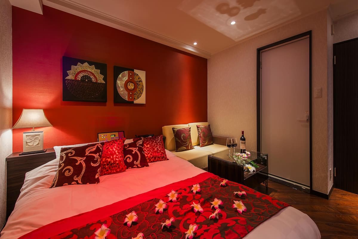 Une chambre d’hôtel avec un grand lit, une table de chevet avec une lampe, deux œuvres d’art murales, une petite table avec une bouteille de vin et des verres, et une porte.
