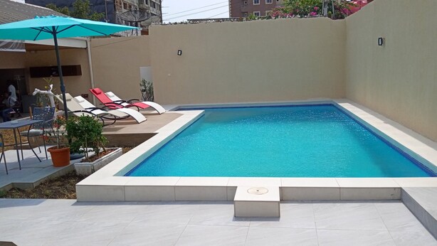 A rectangular swimming pool with a tiled deck, surrounded by a low wall. There are lounge chairs and a table with chairs, and a potted plant near the pool. - Residence Mturquoise T1 (Abidjan)