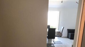 Een smalle gang met een keuken aan de linkerkant, een eethoek met een tafel en stoelen in het midden, en een donkere bank op de achtergrond.