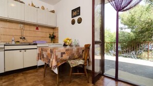 Een keuken met witte kastjes, een houten tafel met een geruit tafelkleed, en een stoel. Er is een raam met uitzicht op bomen en een balkon met een paars gordijn.