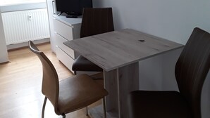 A modern, minimalistic room with a wooden dining table, two chairs, a television, and a window with blinds. - Apartments Sachsenallee (Chemnitz)