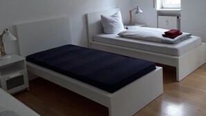 A minimalist bedroom with a single bed, a nightstand, a small table, and a window. - Apartments Sachsenallee (Chemnitz)