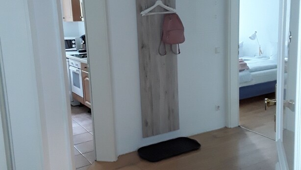 A hallway with a wooden wall, a hanging coat, and a black mat on the floor. - Apartments Sachsenallee (Chemnitz)