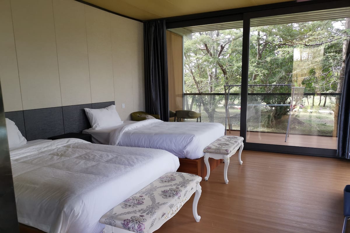 Habitación de hotel con dos camas, un banco y un amplio ventanal que ofrece vistas a árboles y un muro de piedra.