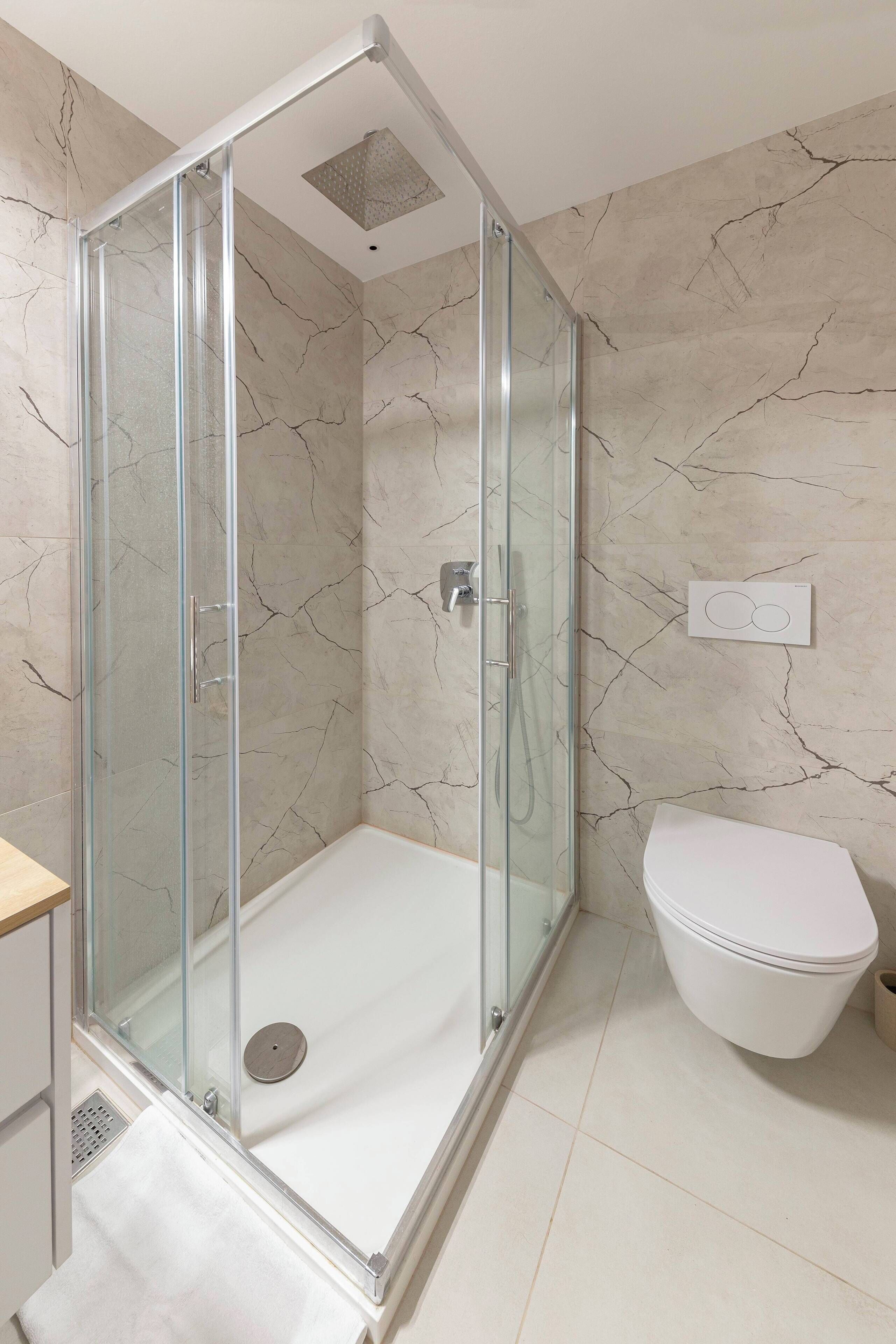 Et moderne badeværelse med et glasvægshus, et hvidt toilet og vægge med marmorlook.