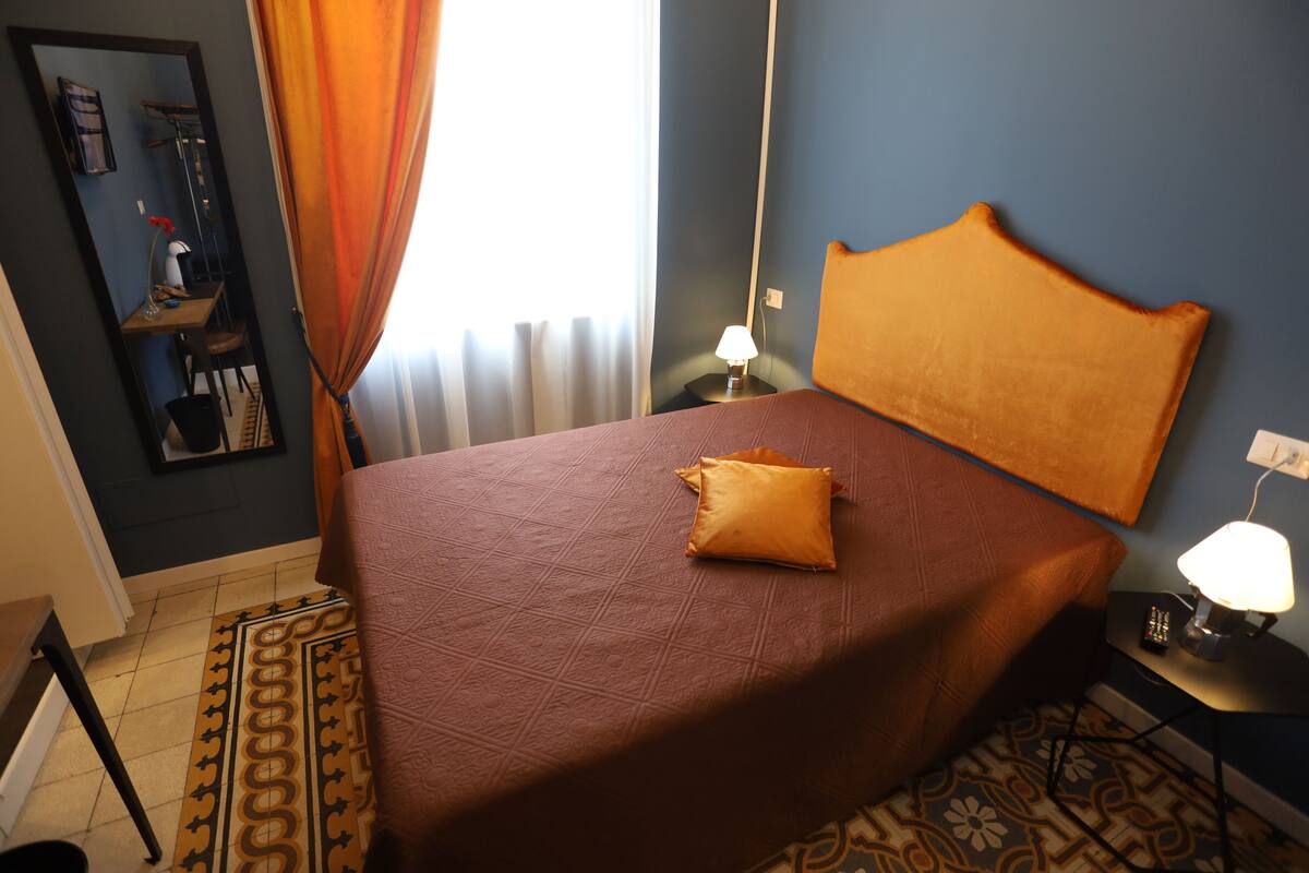 Habitación de hotel con cama, mesita de noche con lámpara, espejo y ventana con cortinas.