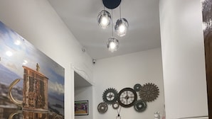 Un estrecho pasillo con suelo de patrón geométrico blanco y negro, un taburete verde, un reloj en la pared y una lámpara en el techo.