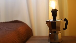 Una hornilla portátil sobre una mesa con una bombilla encendida encima, una cama con cubrecama marrón y una ventana con cortinas.