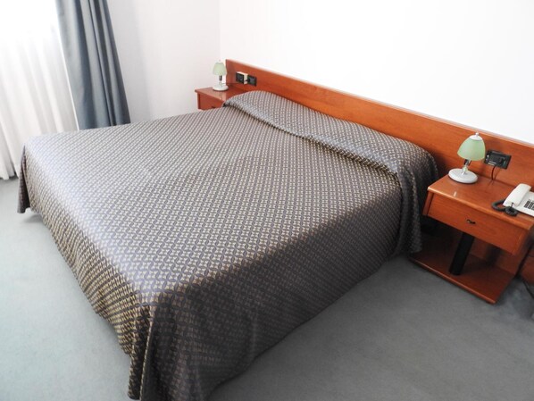 A neatly made bed with a patterned bedspread and pillows, a bedside table with a lamp and a phone, and a window with curtains.