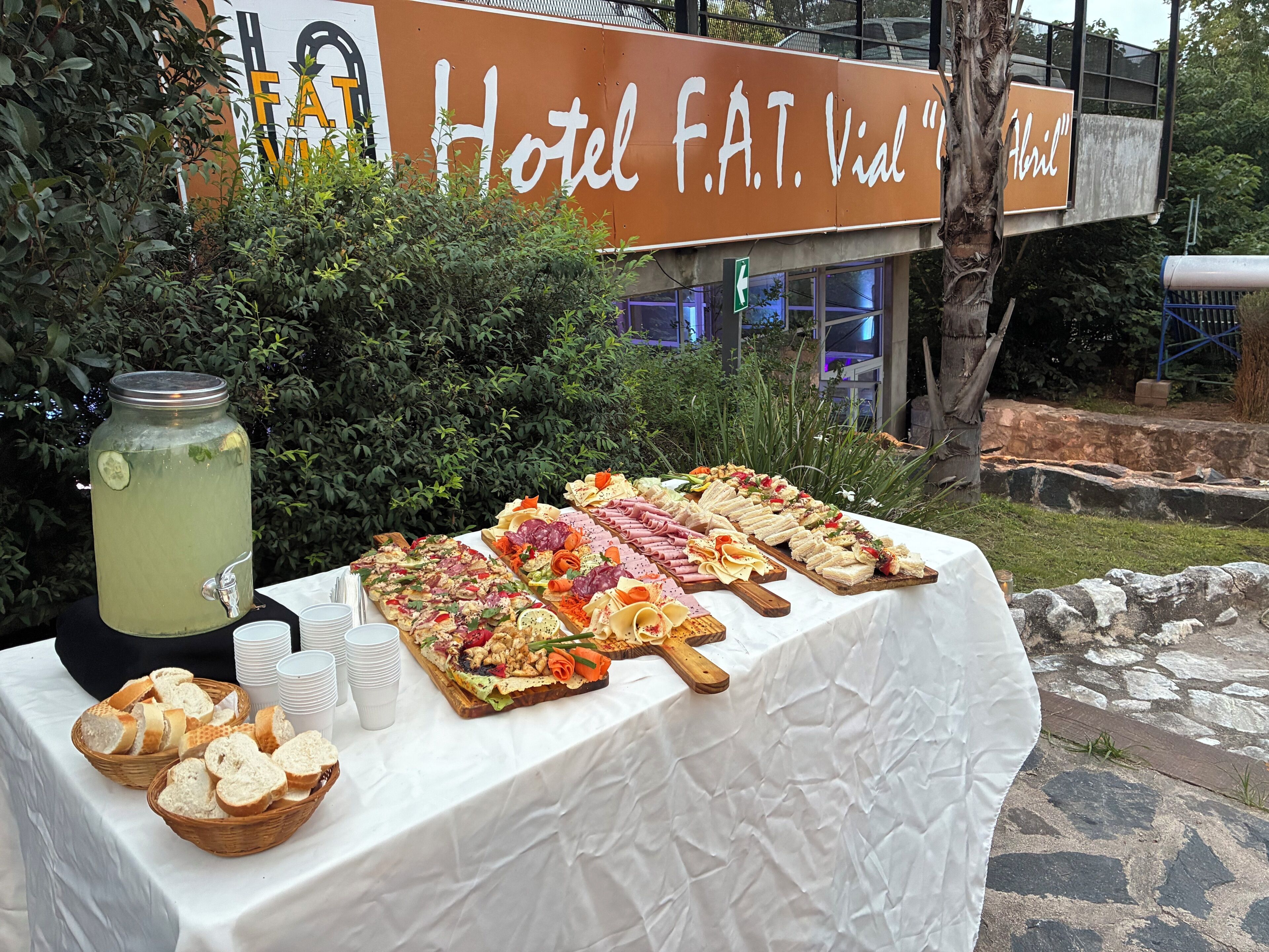 A buffet table with various food items and a beverage dispenser, set outdoors with a hotel sign in the background.