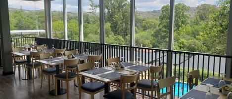 A dining area with tables and chairs, large windows with a view of greenery and hills.