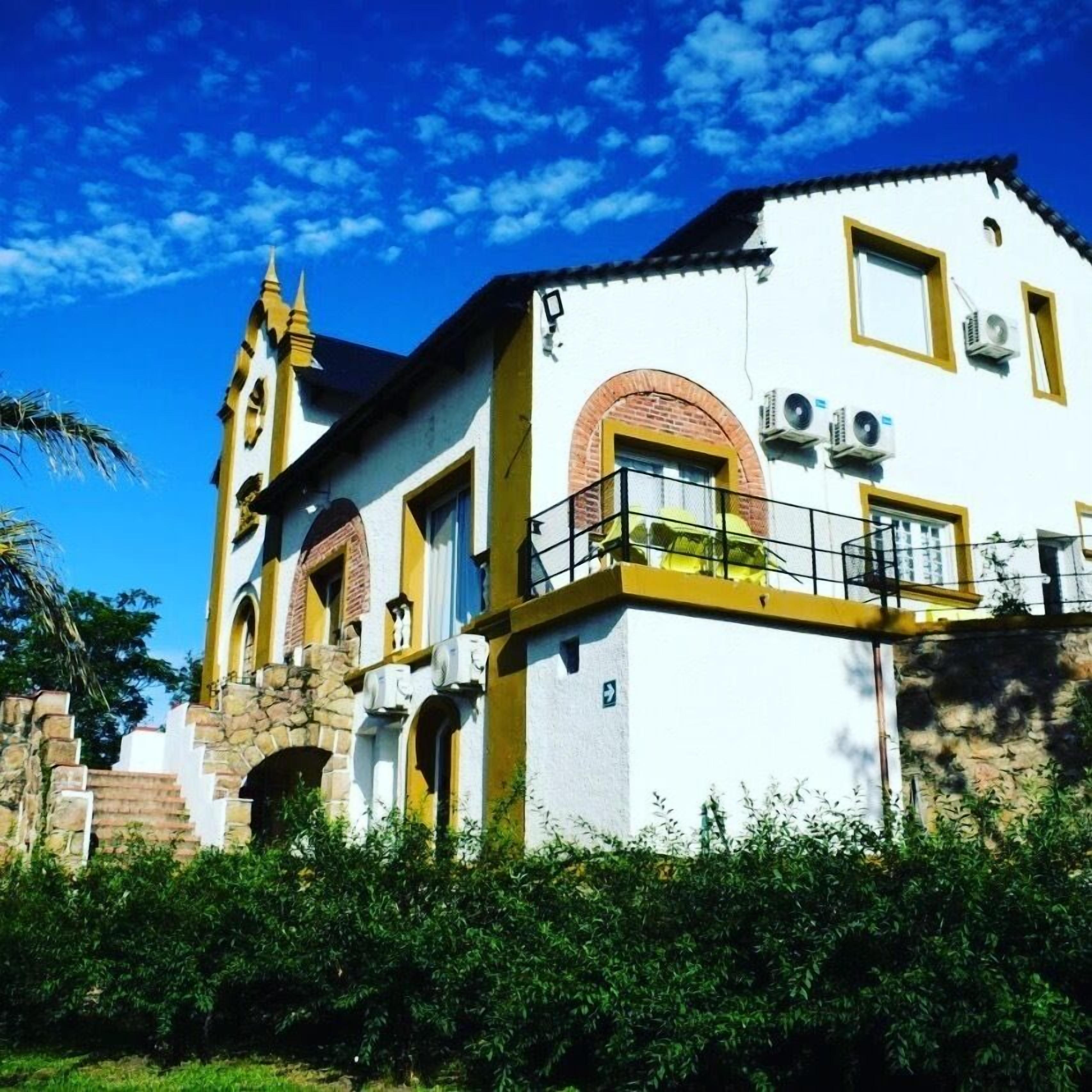A white building with yellow trim and a balcony, surrounded by greenery.