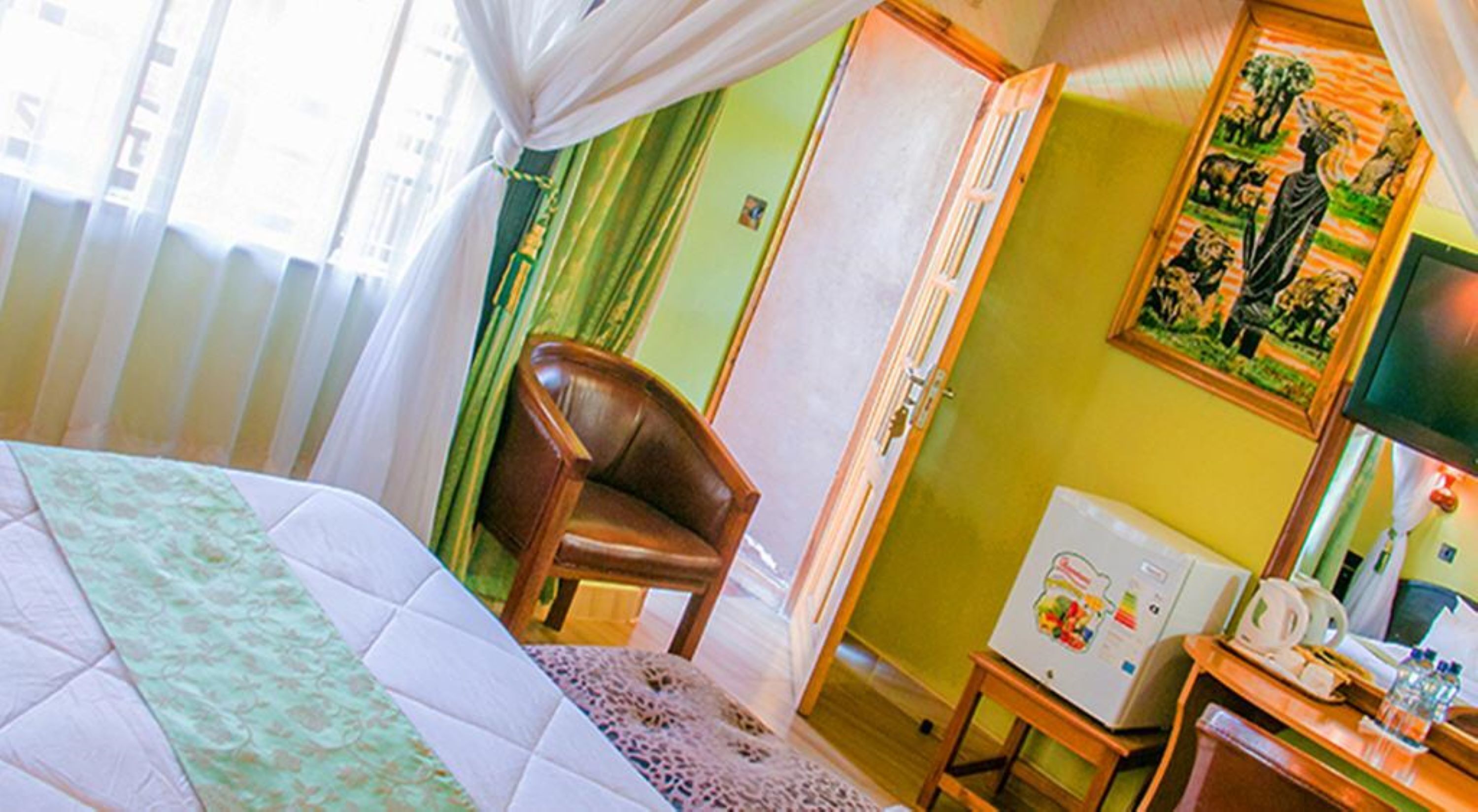 A hotel room with a bed, a chair, a desk with a computer, a refrigerator, and a painting on the wall.