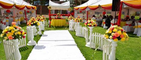Un lugar para bodas con un pasillo blanco, cercas blancas en forma de empalizada y toldos decorados con telas amarillas y rojas.