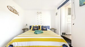 Une chambre avec une couvre-lit Ă rayures, un panneau dĂ©coratif en forme de poisson accrochĂ© au mur, une petite table de chevet avec une lampe et une fenĂȘtre donnant sur lâextĂ©rieur.