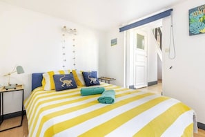 A bedroom with a bed featuring yellow and white striped bedding, two pillows with monkey designs, a teal towel, and a nightstand with a lamp.