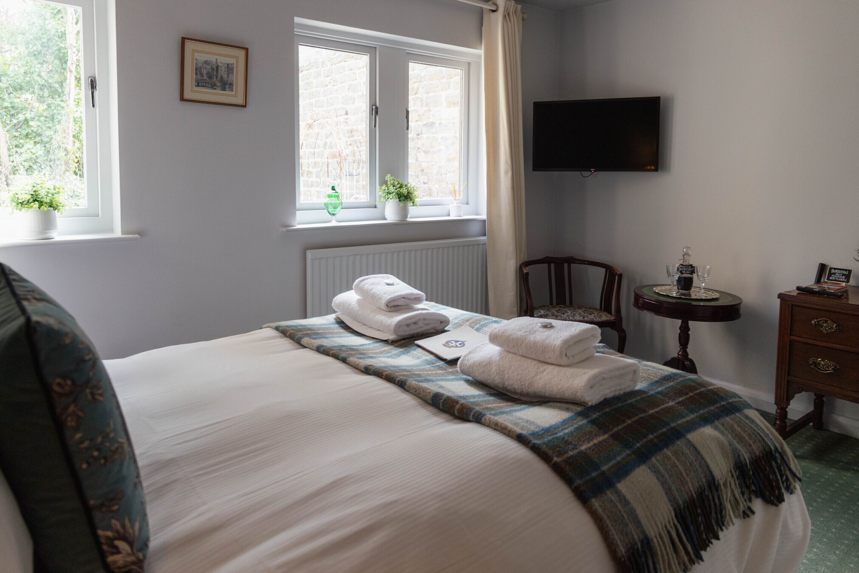 A neatly made bed with towels, a bedside table with a bottle and glass, a chair, a small table with a remote control, and a TV on the wall.