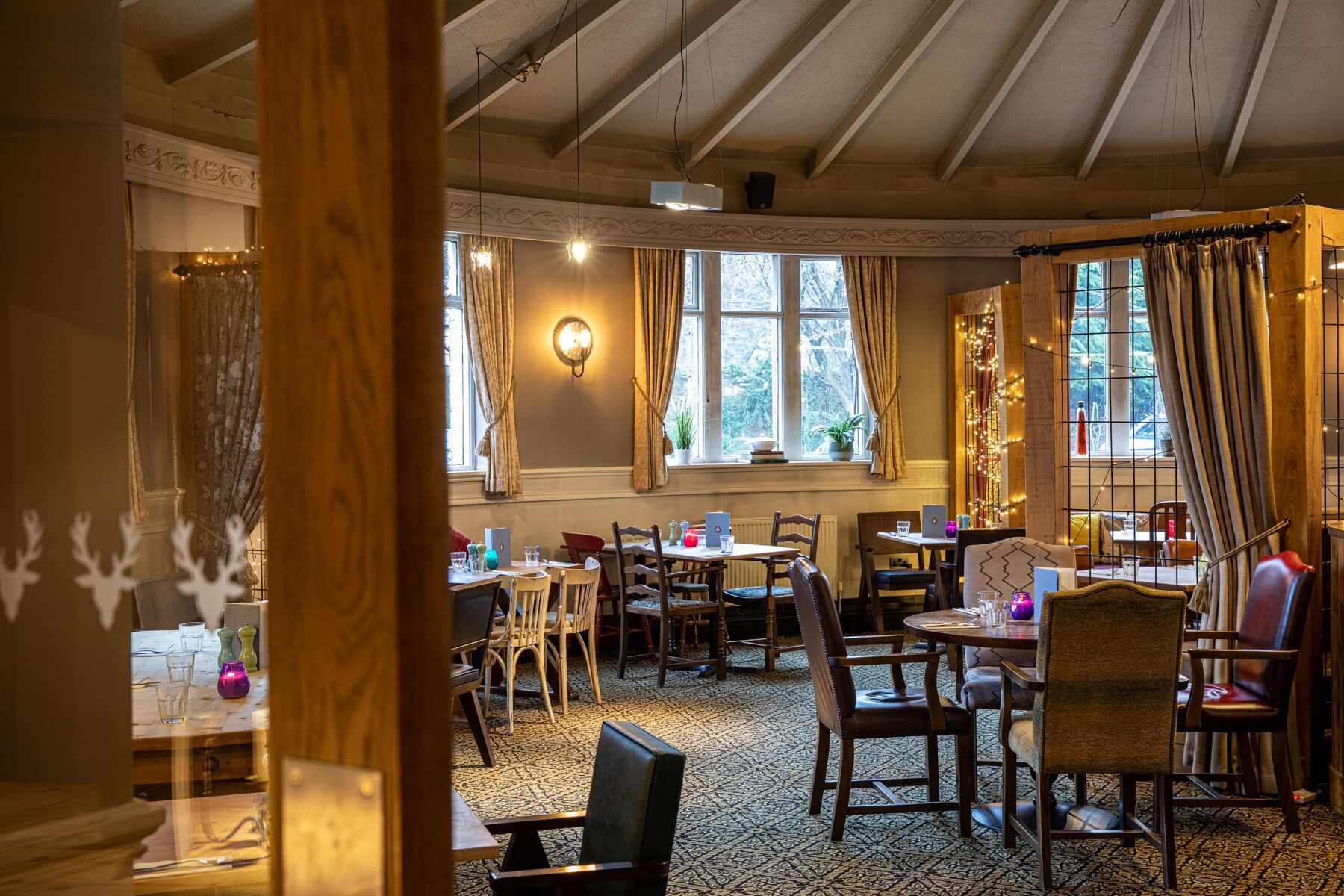 A cozy dining area with wooden tables and chairs, large windows with curtains, and a warm ambiance.