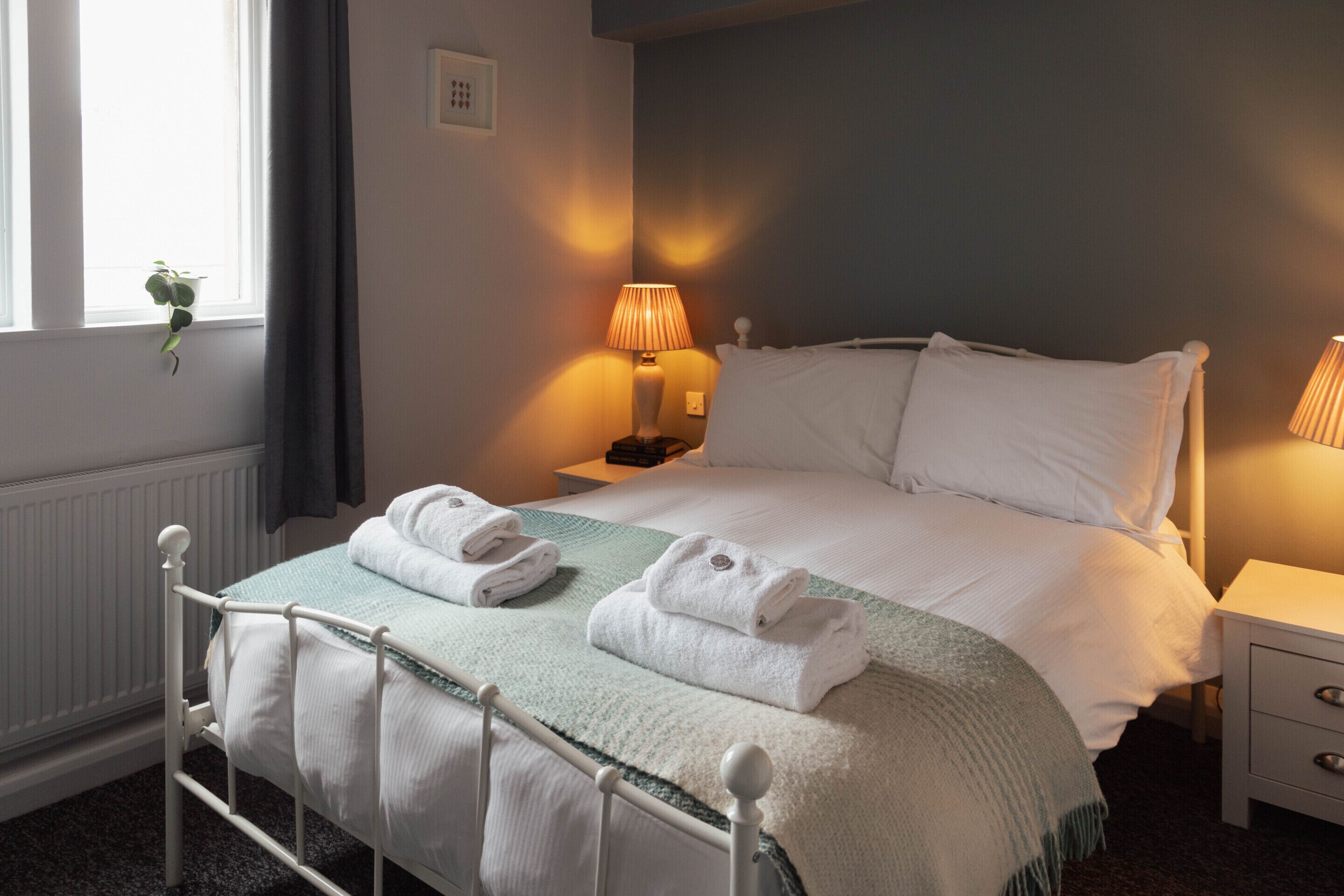 A neatly made bed with white linens and a green throw blanket, a bedside lamp, and a window with a plant.