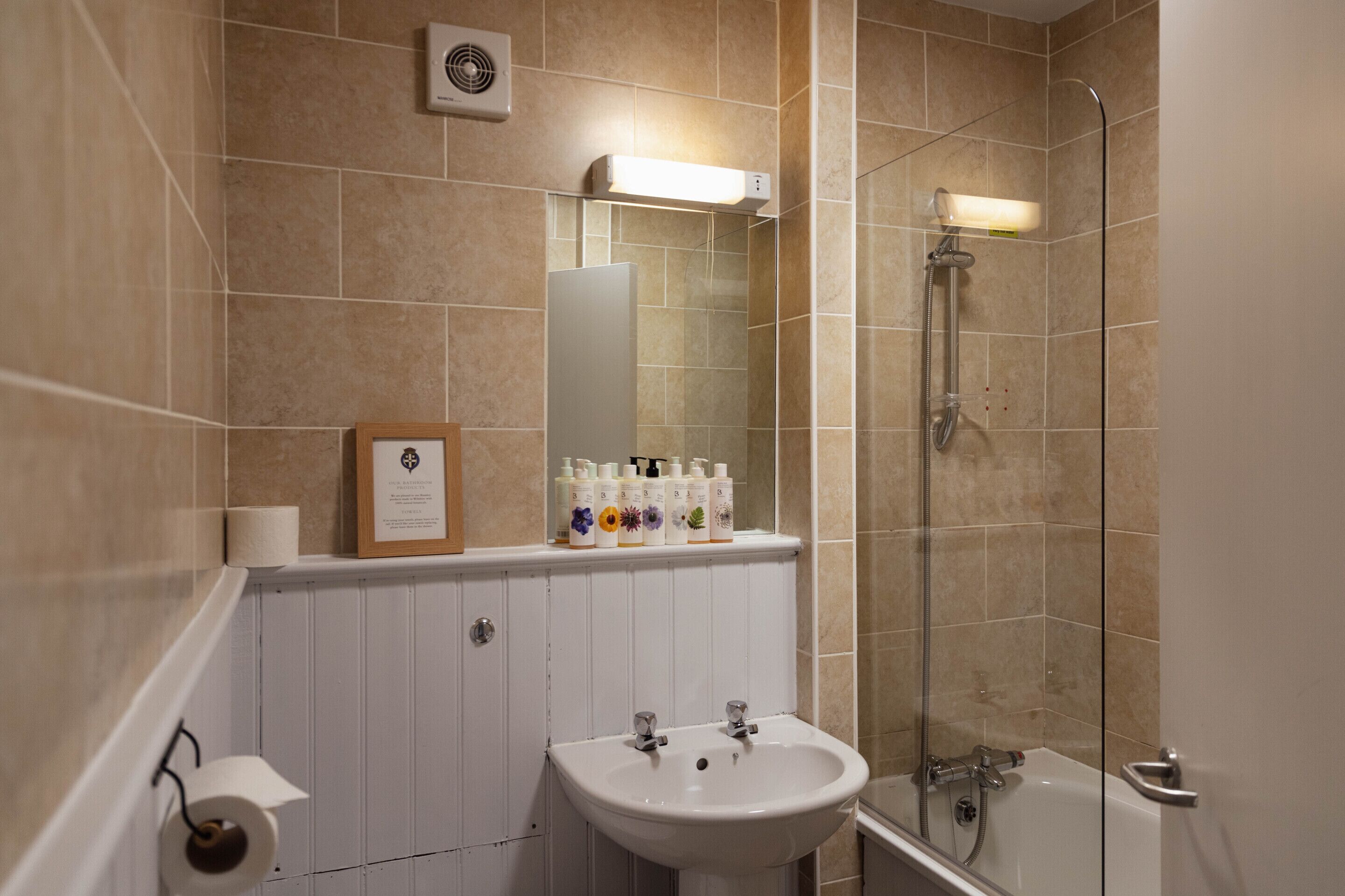 A bathroom with a sink, mirror, shower, and toilet paper.