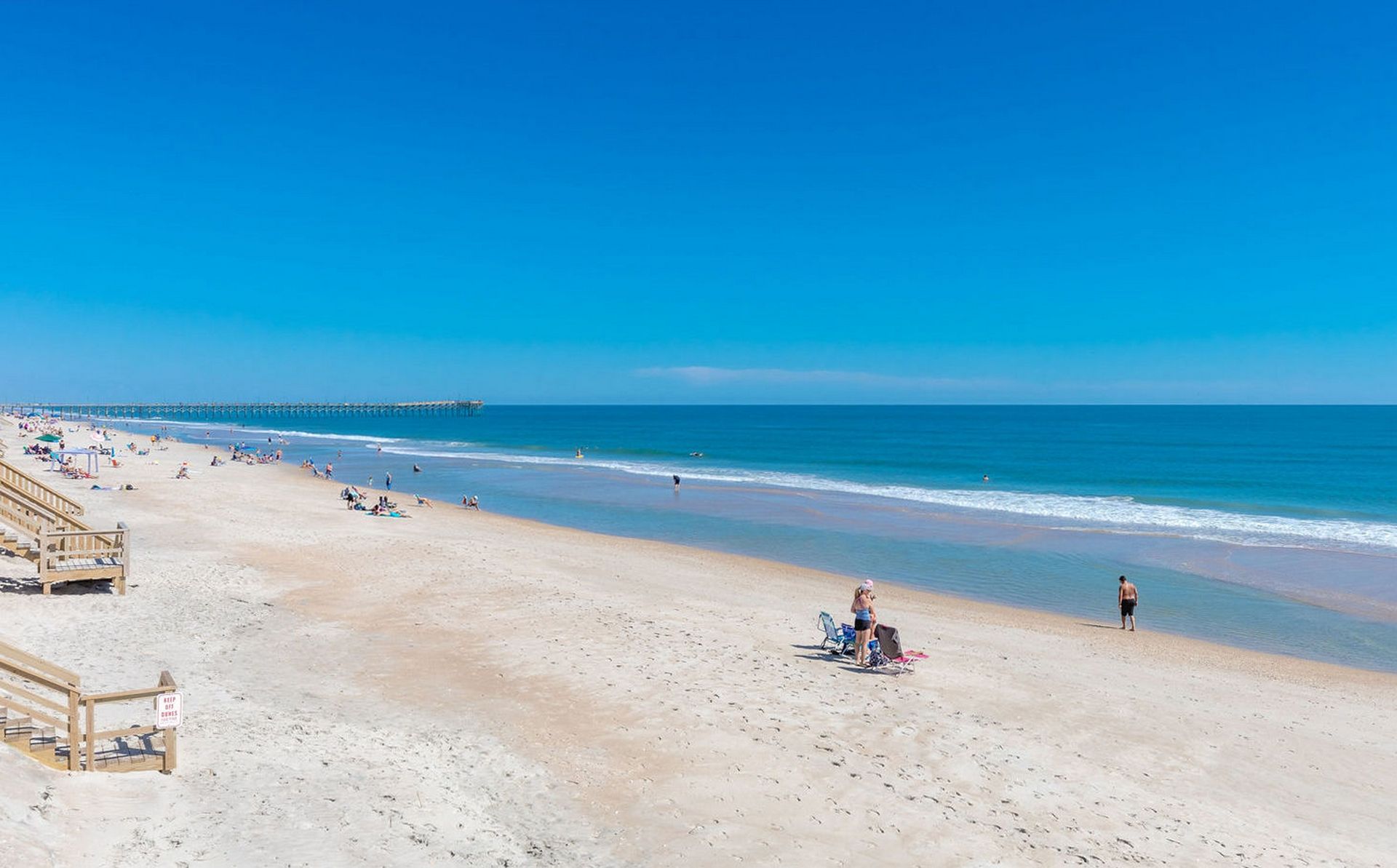 A beach with a wooden walkway leading to the water, a pier in the distance, and people relaxing on the sand.