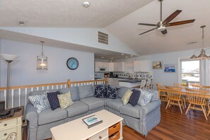 A modern living room with a sectional sofa, a dining area with wooden chairs, a kitchen in the background, and a ceiling fan. - Topsail State of Mind - 4BR Oceanfront Home; Elevator; Covered Porch (Surf City)