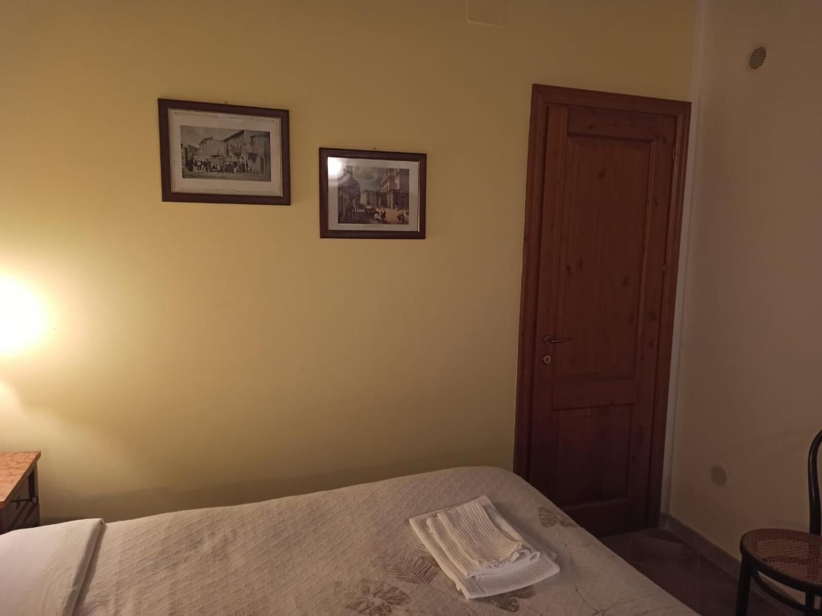 Ein Zimmer mit einem Bett, zwei gerahmten Bildern an der Wand, einer Holztür und einem Stuhl.