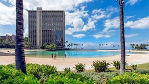 Hotel frente a la playa con una piscina grande, palmeras y un cielo azul despejado.