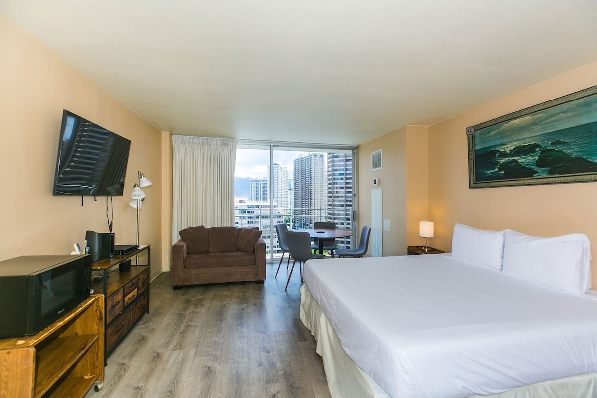 A hotel room with a bed, a TV, a desk with a chair, a couch, and a view of high-rise buildings.
