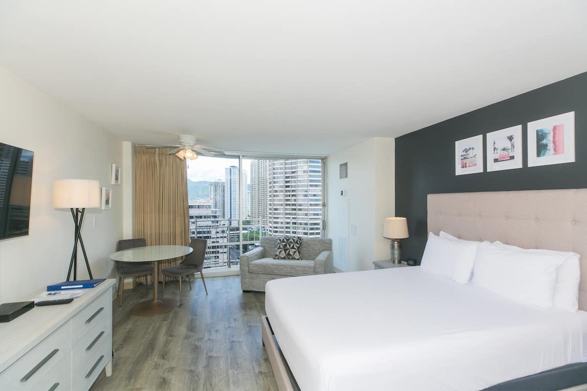 A hotel room with a large bed, a desk, a chair, a small table, and a view of high-rise buildings.