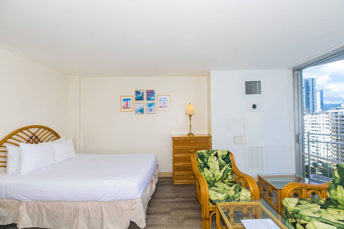 A hotel room with a bed, a desk with a chair, a lamp, a dresser, and a large window offering a city view.
