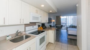A modern kitchen with white cabinets, a microwave, stove, and sink. There is a dining area with a table and chairs, and a view of a cityscape through large windows. - Ilikai Marina Studio City View Condos with Fully Equipped Kitchens & Free Wifi (Honolulu)