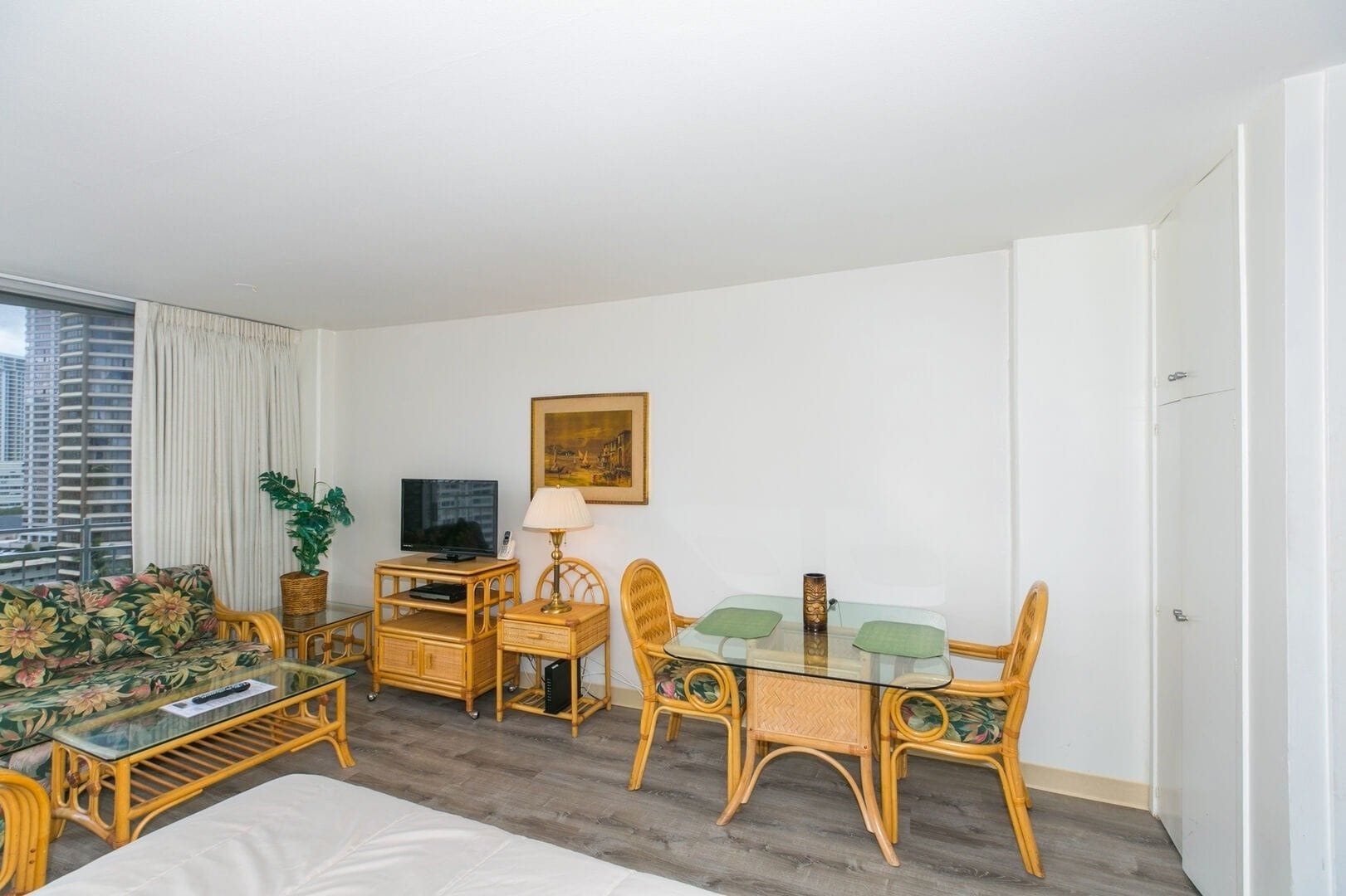A hotel room with a bed, a television, a dining table with chairs, a lamp, and a window with a view of buildings.