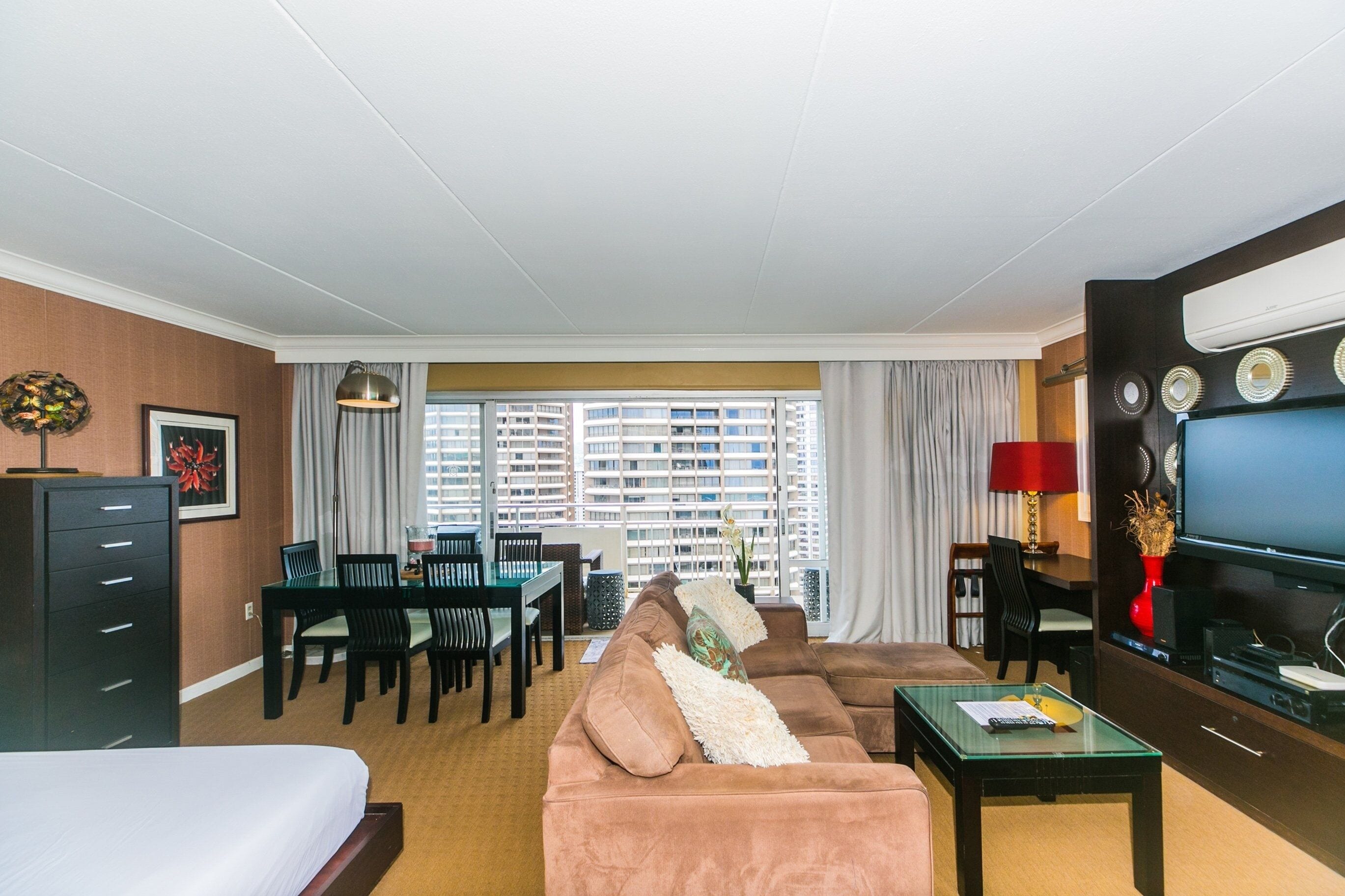 Une chambre d’hôtel avec un grand lit, un canapé, un coin repas avec des chaises, une télévision et une vue sur les gratte-ciel.