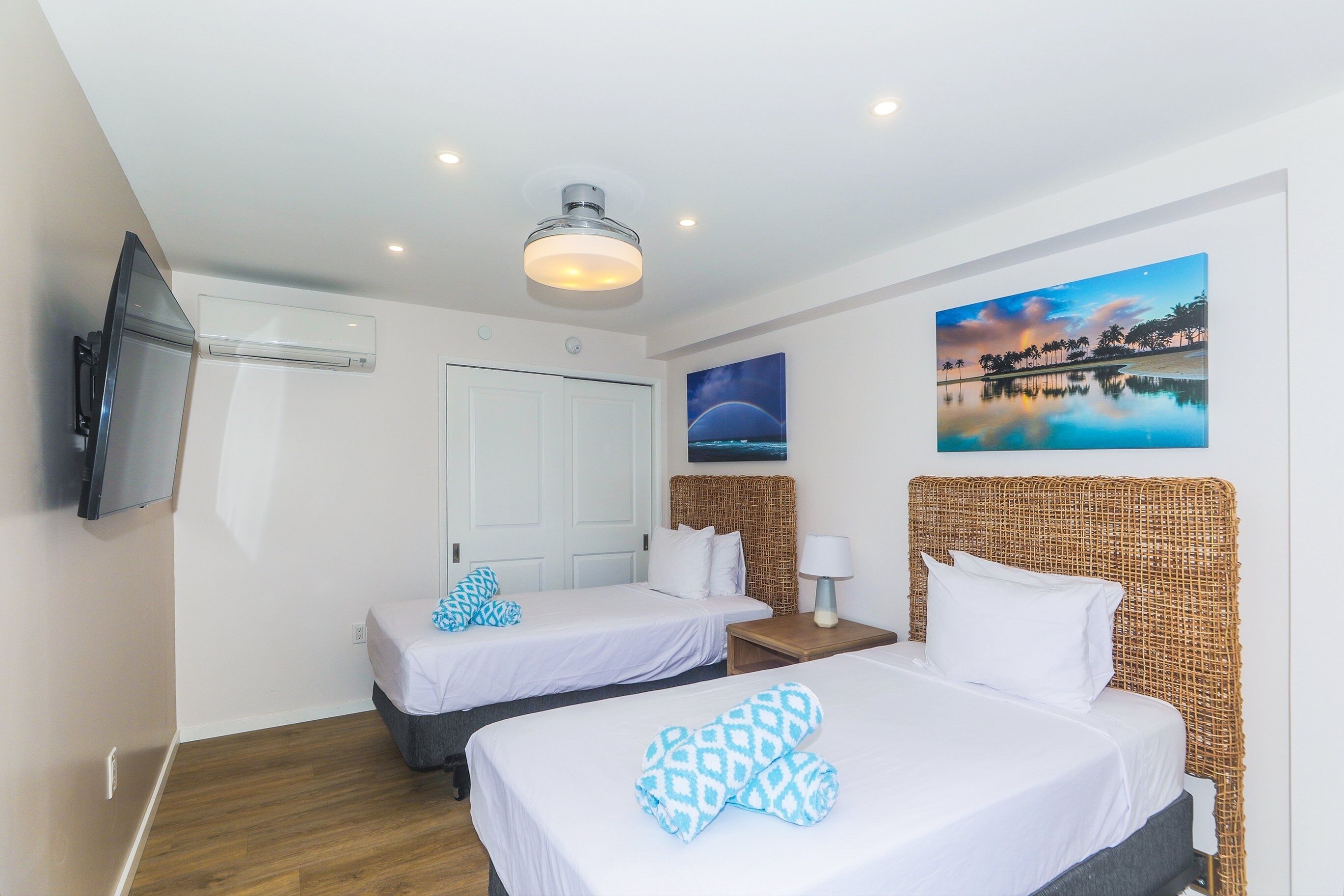 A hotel room with two beds, a television, a painting, and a ceiling fan.