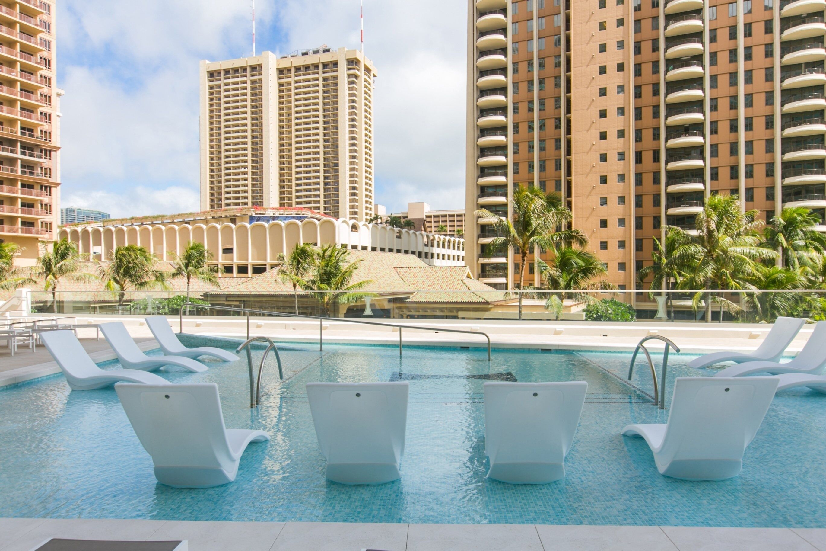 Una piscina con sillas reclinables blancas, rodeada de palmeras y edificios altos.