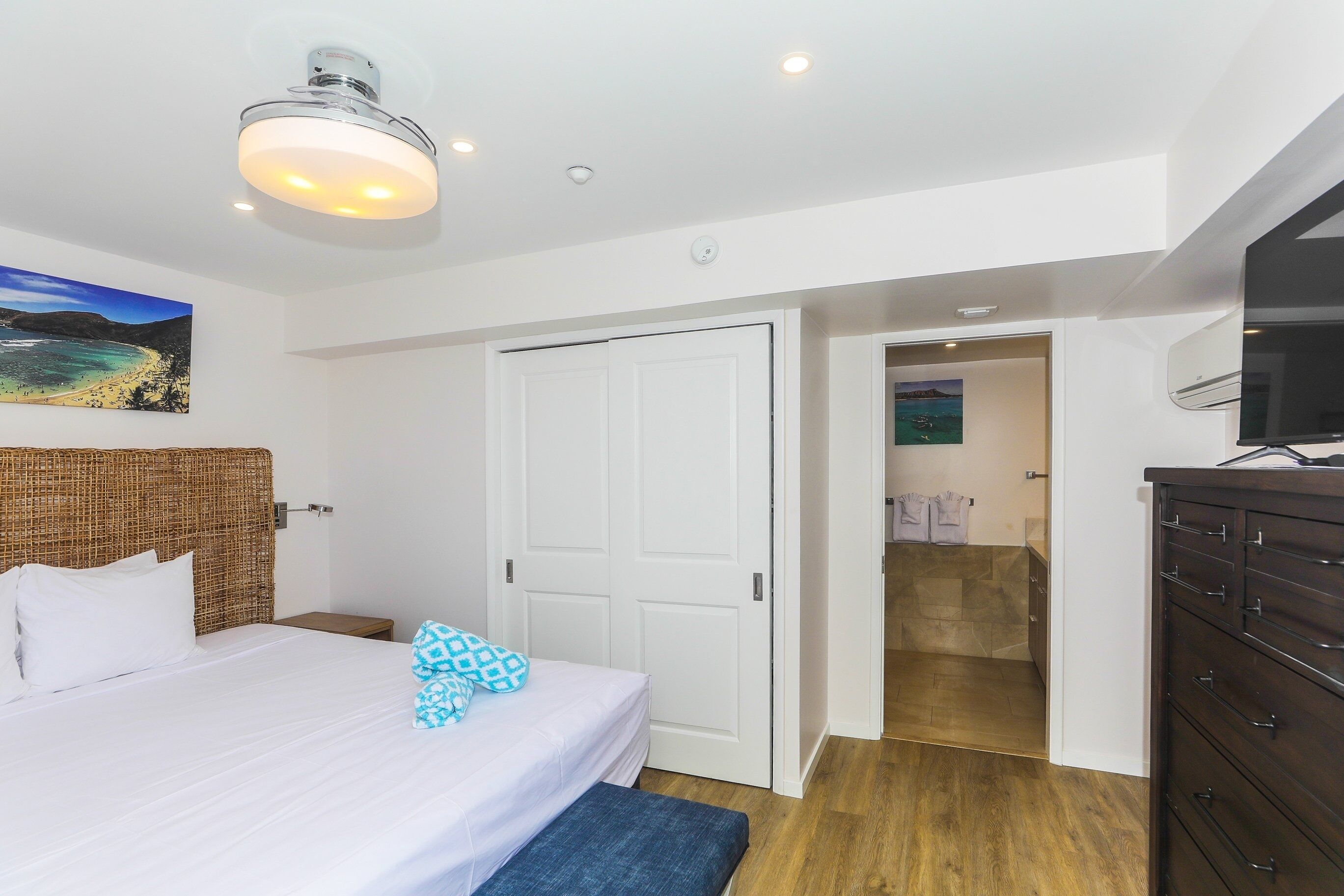Un dormitorio con una cama, una puerta blanca, un banco de madera, un aparador y un cuadro en la pared.