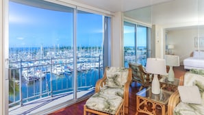 A spacious room with a large sliding glass door leading to a marina view. Inside, there is a wooden chair, a glass coffee table, and a white sofa. - Ilikai Marina One Bedroom Harbor View Condos - Walk to Ala Moana Beach Park! (Honolulu)